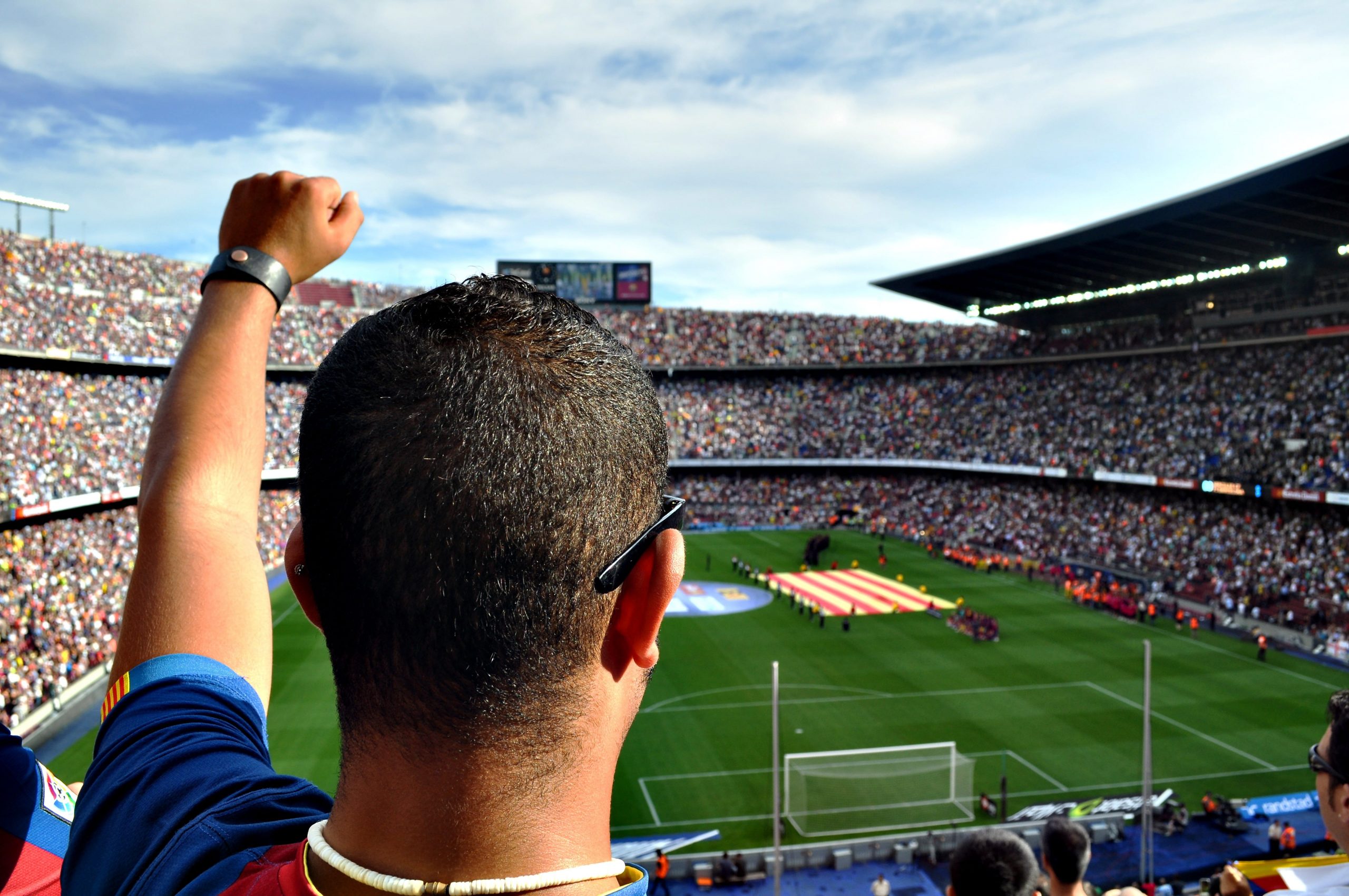 A man with short, dark hair and his fist raised in the air stand in the foreground in front of a full soccer stadium with the field in the background.