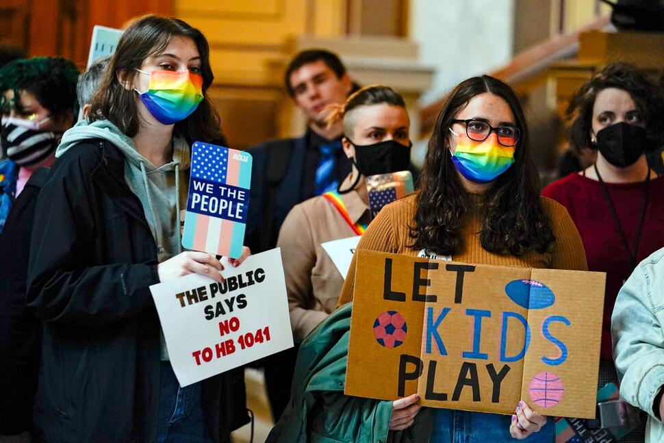 People wearing ranbow colored masks hold signs reading, "let kids play" and "the public says no to HB 1041"