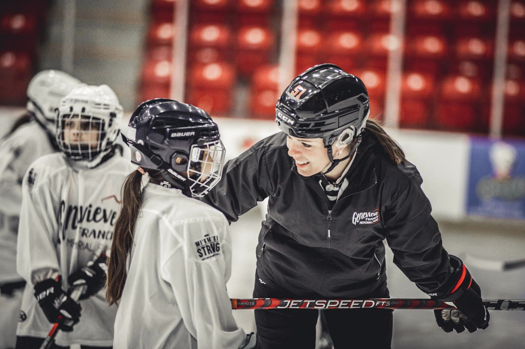 An ice hockey coach, dressed in black, leans over on the ice, holding her stick across her legs, talking to a young participant with a long pony tail and wearing a white jersey, with other participants standing in the background.